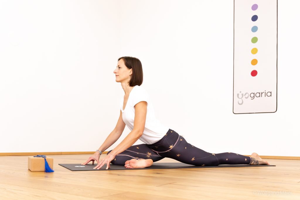 Klaudia, die Eigentümerin der Yogaria in der Position der Taube, man sieht sie von der Seite im großen Yogaraum, vor ihr steht ein Block mit einem Yogamala und im Hintergrund sieht man ein Wandbild mit dem Logo der Yogaria und den 7 Punkten der 7 Chakrafarben