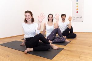 Elli, Klaudia und Christoph sitzen im Hauptraum der Yogaria am Boden und twisten über ihre Knie zur rechten Seite. Ihre Handflächen zeigen nach oben als Zeichen der Begrüssung. Im Hintergrund sieht man das Bild mit Yogaria Logo und den 7 Chakren-Farben
