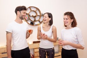 3 Personen im Yogaraum, sie trinken Tee, unterhalten sich und lachen aus vollem Herzen.