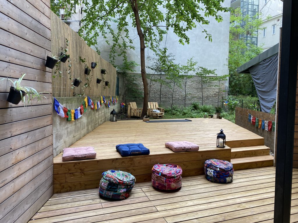 Yogadeck im Garten der Yogaria mit bunten Sitzkissen, 2 Laternen, Nepalfahnen an den Mauern, am Ende des Yogadecks sieht man eine Yogamatte und dahinter noch 2 Gartenmöbel aus Paletten gebaut, ein Blick ins Grüne