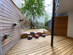 Blick in den Yogaria Garten auf das große neue Yogadeck aus Holz, bunte Sitzkissen, 2 Laternen und der Blick ins Grüne