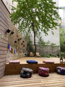 Yogadeck aus der Nähe mit Zoom auf die Sitzkissten und der große Baum im Zentrum des Gartens thront über allem