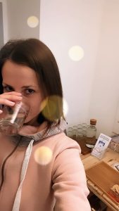 Wasserfilter Klaudia trinkt ein Glas gefiltertes Wasser und im Hintergrund sieht man die Informationsbroschüren