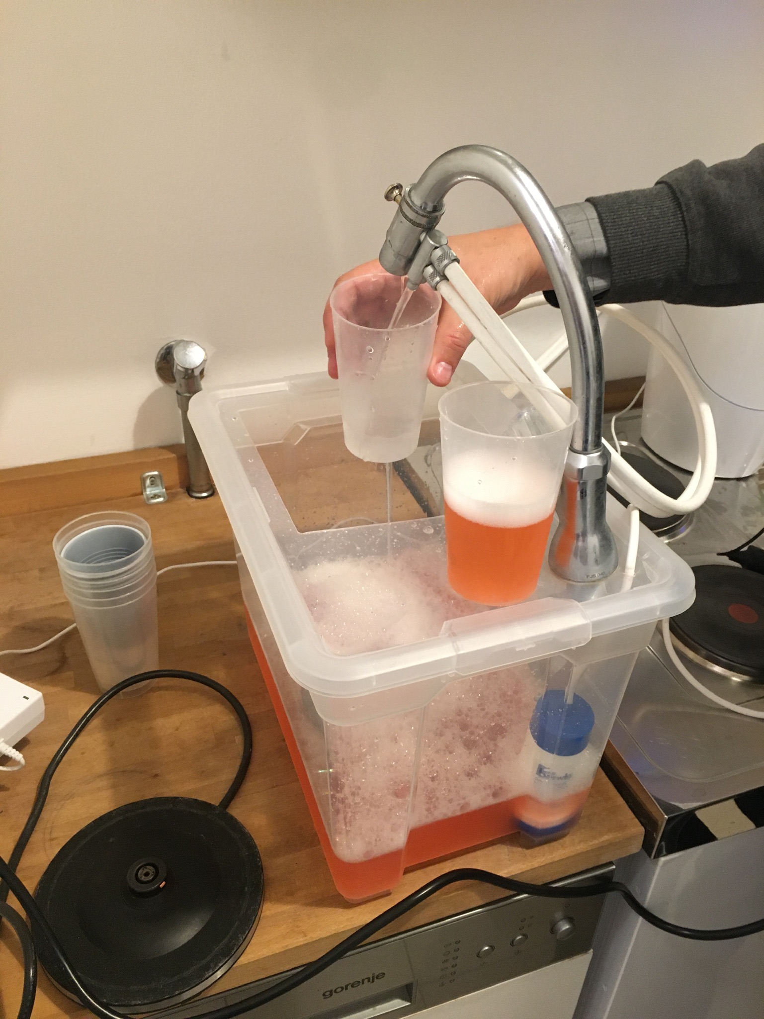 man sieht einen Behälter mit orangener Flüssigkeit, 2 Becher, 2 Schläuche und das gefärbte Wasser kommt am anderen Ende gefiltert und klar heraus.