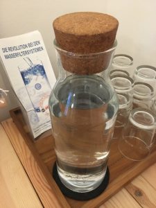 ein Glaskrug mit Wasser gefüllt, steht auf einem Holzbrett vor leeren Gläsern und vor einem Ständer mit Infobroschüren auf denen steht: "Die Revolution bei den Wasserfiltersystemen, eSpring"