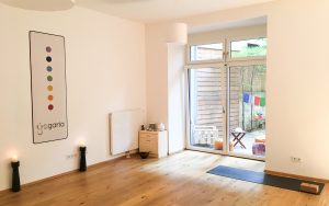 der Yogaraum der Yogaria mit einer Yogamatte am Boden und einem Yogablock darauf, 2 Kerzenständer und der Blick in den Garten über die geschlossene Terrassentür