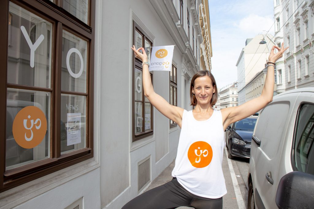 Klaudia steht vor dem Gebäude der Yogaria in der Haltung des Baumes, beide Hände Richtung Himmel gestreckt, sie lächelt, im Hintergrund sieht man die Fenster straßenseitig der Yogaria samt der Yogaria Fahne