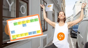 Online Live Yogastunen Klaudia steht in der Postition des Baumes auf dem Gehsteig vor der Yogaria in der Lederergasse, links im Bild ist ein Laptop abgebildet, auf dem man grob einen Stundenplan erkennen kann, die Kästchen haben die Farben Gelb, Grün und Blau, im Hintergrund sieht man die Yogaria Fahne