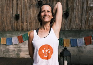 Klaudia im Fersensitz auf ihrer lila Yogmatte mit einem Yogaria Tshirt im Garten auf dem Yogadeck der Yogaria mit einer Schulterdehnung und einem heiteren Gesichtsausdruck
