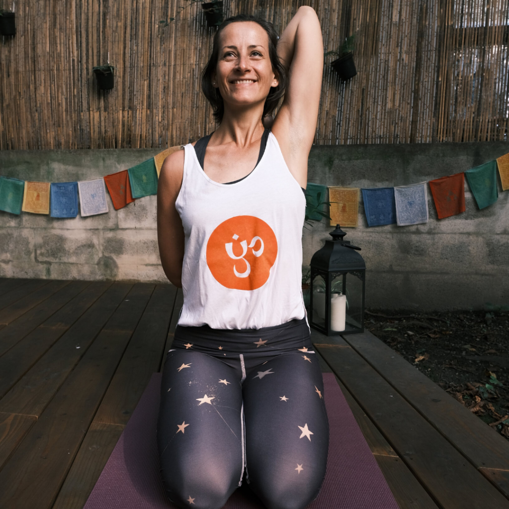 Klaudia im Fersensitz auf ihrer lila Yogmatte mit einem Yogaria Tshirt im Garten auf dem Yogadeck der Yogaria mit einer Schulterdehnung und einem heiteren Gesichtsausdruck