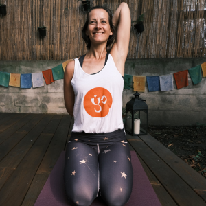 Klaudia im Fersensitz auf ihrer lila Yogmatte mit einem Yogaria Tshirt im Garten auf dem Yogadeck der Yogaria mit einer Schulterdehnung und einem heiteren Gesichtsausdruck
