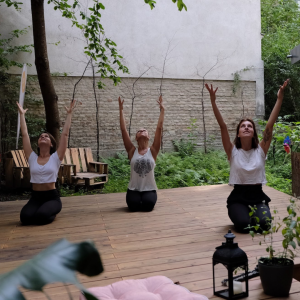3 Yogis sitzen im Fersensitz auf dem Yogadeck im Garten der Yogaria, sie haben die Augen geschlossen und strecken ihre Arme zum Himmel, im Vordergrund sieht man eine Laterne, einen rosaroten Polster und ein paar Pflanzen ragen ins Bild, im Hintergrund ist der Garten der Yogaria zu sehen
