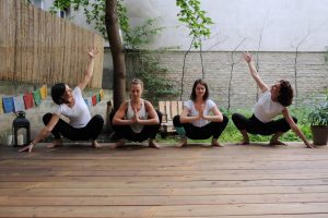 auf dem Yogadeck im Garten der Yogaria, 4 Yogalehrerinnen in der Position von Malasana, in der Hocke, Ellbögen gegen die Innenseite der Knie gedrückt, lächelnd