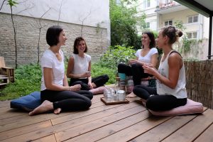 4 Yogalehrerinnen sitzen im Kreis auf dem Yogadeck im Garten der Yogaria und unterhalten sich, sie trinken Tee und Wasser und sprechen miteinander