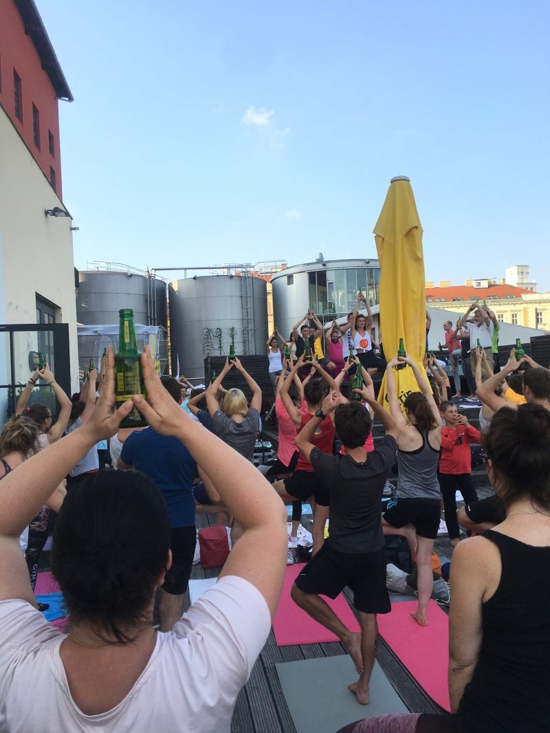 Unzählige Menschen beim Yoga im Freien, alle stehen in der Position des Baums und balancieren eine Bierflasche auf dem Kopf, im Hintergrund sieht man große Biertanks und gelbe Sonnenschirme