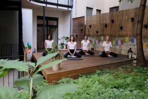 4 Yogalehrerinnen im Schneidersitz auf der Terrasse der Yogaria, sie haben die Augen geschlossen, die Hände im Schloss gefaltet und die Augen geschlossen, sie meditieren, im Vordergrund viel grün des Gartens