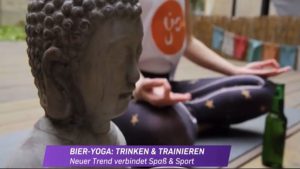 im Vordergund der Kopf einer kleinen Buddhastatue in der Yogaria, im Hintergrund Klaudia im Lotussitz, meditativ, mit einer Bierflasche vor ihr auf dem Boden