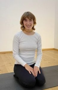 Bella im Heldensitz lächelt in die Kamera, auf der schwarzen Matte in der Yogaria