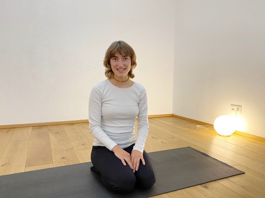 Bella Tröstl im Porträt, sitzend auf der schwarzen Yogamatte in der Yogaria, sie lächelt in die Kamera