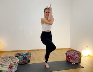Kathi Wabnigg in der Haltung des Adlers, sie steht auf einer schwarzen Yogamatte mit verschränkten Armen und gekreuzten Beinen und lächelt in die Kamera