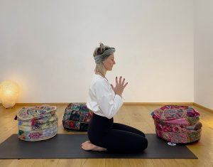 Kathi in einer meditativen Haltung im Fersensitz von der Seite, die Hände zur Gebetshaltung gefaltet und die Augen geschlossen, sie sitzt auf einer schwarzen Yogamatte am Boden