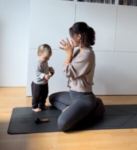 Mama und Baby sitzend auf einer Yogamatte, die Mama hat ihre Hände zu Namaskar gefaltet und blickt zu ihrem Kind, das Kind steht vor der Mama und blickt zu Boden