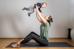 Klaudia sitzt auf ihrer schwarzen Yogamatte, sie streckt ihre Hände zur Decke und hält ein Baby in ihren Händen, beide lachen