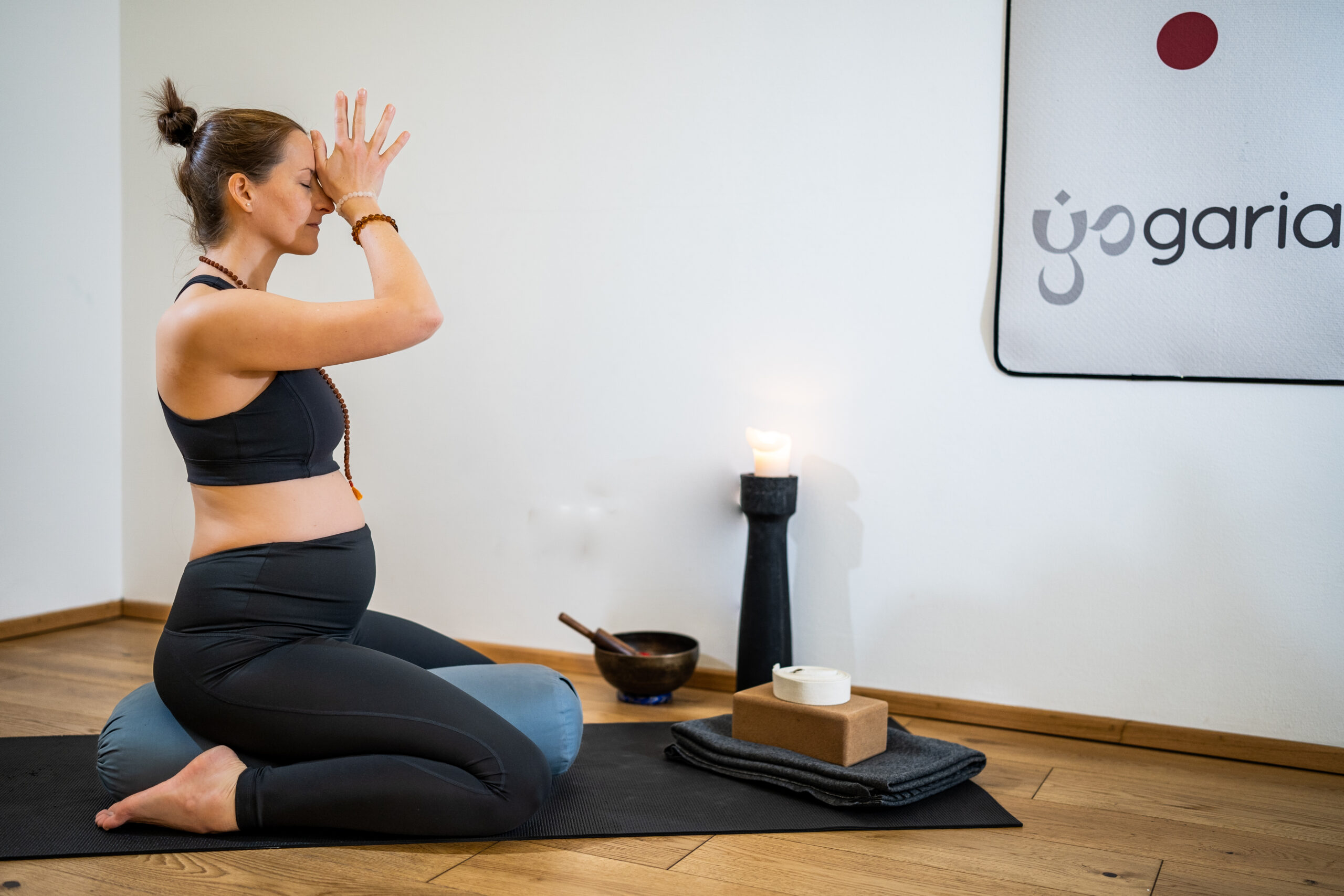 Klaudia ist schwanger und sitzt im Fersensitz auf einem Yogabolster und hat die Hände meditativ vor ihrem 3. Auge gefaltet, man sieht sie von der Seite und sie ist schwanger