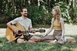 Marie & Paul Paul und Marie sitzen in der Wiese, Paul spiel Gitarre und lächelt, Marie lächelt zurück