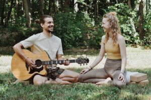 Paul und Marie sitzen in der Wiese, Paul spiel Gitarre und lächelt, Marie lächelt zurück