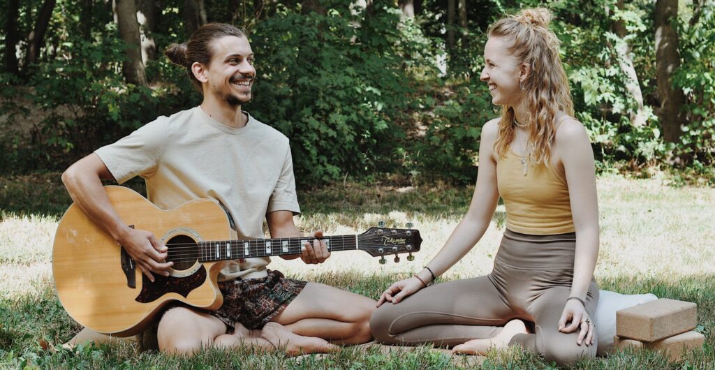 Paul und Marie sitzen in der Wiese, Paul spiel Gitarre und lächelt, Marie lächelt zurück