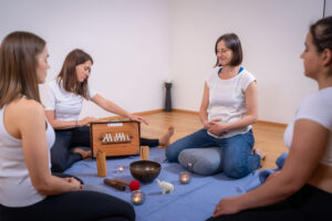 Yogaria Team, 4 Frauen sitzen im Kreis auf bequemen Polstern, eine Frau spielt das Harmonium und die anderen hören entspannt zu